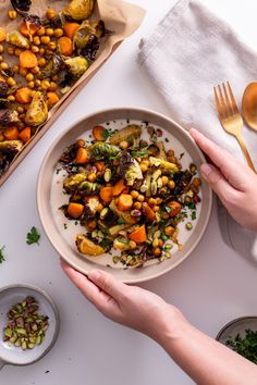 two hands holding a plate with roasted vegetables on it, next to a baking dish