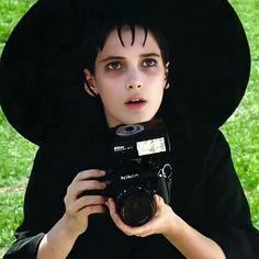 a young boy dressed in black holding a camera and taking a photo with his hat on