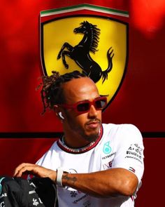 a man with dreadlocks standing in front of a red ferrari logo and wearing sunglasses