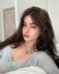 a woman with long brown hair sitting on a bed wearing a gray shirt and silver necklace