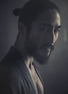 a man with a bun in his hair looks into the distance while standing against a dark background