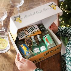 a woman opening a gift box filled with gin tonics and lemonade next to a christmas tree