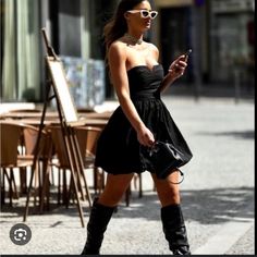 a woman in black dress and knee high boots looking at her cell phone while walking down the street