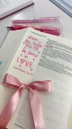 an open book with pink ribbon and writing on it, next to two pens and a pen