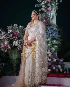 a woman in a white and gold sari is standing on a stage with flowers behind her