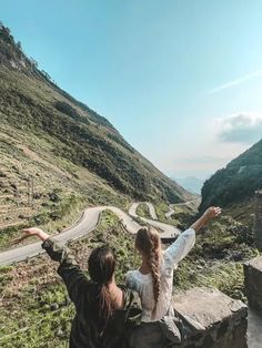 two people sitting on the side of a road with their arms in the air and one person pointing to the sky