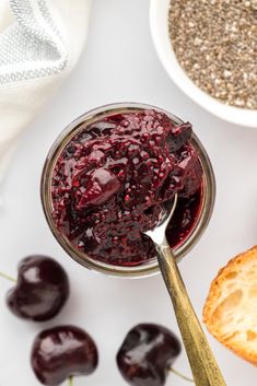 a spoon with jam in it next to some cherries and bread on the table