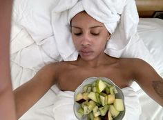 a woman laying in bed with a towel on her head holding a bowl of fruit