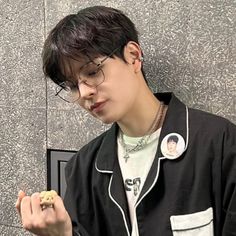 a young man wearing glasses holding a cookie and looking at his cell phone while standing against a wall