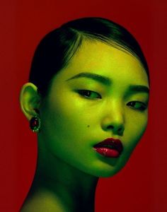 a woman with green makeup and earrings on her head, looking at the camera while standing against a red background