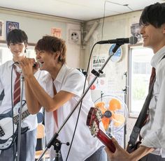 three young men singing into microphones in front of an open mic stand with guitars