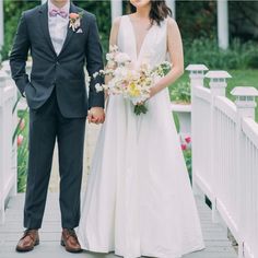 a bride and groom are standing on a bridge holding hands, dressed in black tuxedos