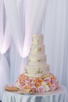 a white wedding cake with pink and yellow flowers on the table in front of sheer drapes