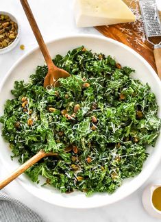 a white bowl filled with greens and nuts on top of a wooden cutting board next to cheese