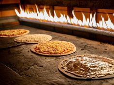 three pizzas sitting in front of an oven with fire coming from the wall behind them