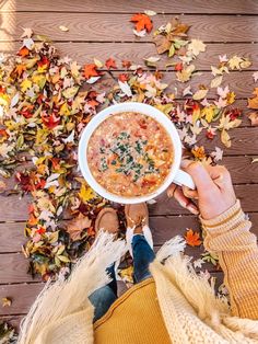 a person holding a bowl of soup in front of autumn leaves on a wooden floor