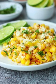 a white plate topped with corn salad next to lime wedges and cucumbers