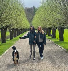 two women walking down a path with a dog in the foreground and trees behind them