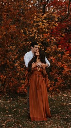 a man and woman standing next to each other in front of trees with orange leaves