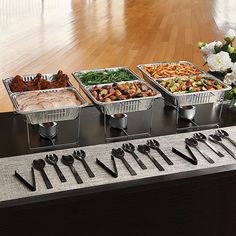 four trays of food on a table with utensils and flowers in the background