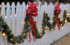 christmas garlands are hung on a white picket fence, decorated with red bows and lights