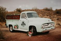 an old white truck with flowers in the bed is parked on dirt near bushes and trees