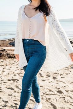 a woman standing on top of a sandy beach next to the ocean with her hands in her pockets