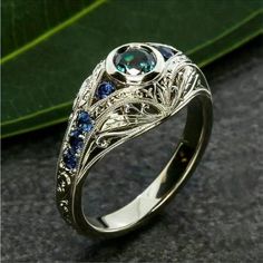 a white gold ring with blue sapphires and diamonds on the side, sitting next to a green leaf