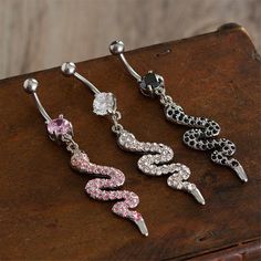 three pairs of dangling belly rings with pink and white crystals in them on a piece of wood