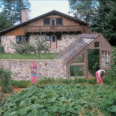 two people in front of a stone house with vegetables growing on the ground and trees behind them