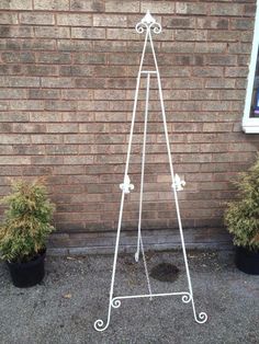 a white metal easel stands in front of a brick wall with potted plants