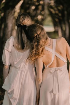 two women in white dresses are walking through the woods with their backs to each other