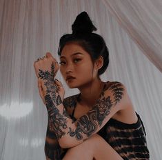 a woman with tattoos on her arm sitting in front of a white curtain and looking off to the side