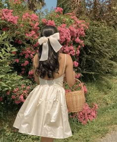 a woman in a white dress is holding a basket and looking at some pink flowers