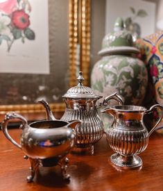 three silver teapots sitting on top of a wooden table next to vases