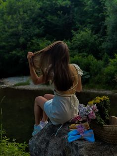 a woman sitting on top of a rock next to a river with flowers in her hair