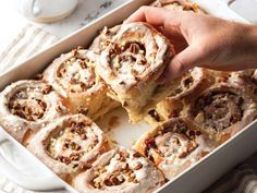 a person holding a piece of cinnamon roll in a white casserole dish with pecans on the side