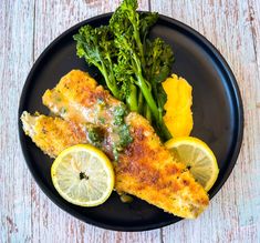 a black plate topped with fish, lemon wedges and broccoli on top of a wooden table