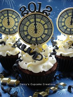 three cupcakes with white frosting and black numbers on them are in front of a clock