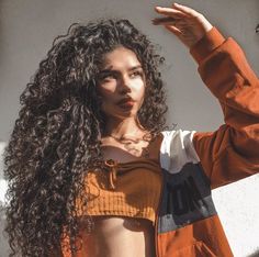 a woman with long curly hair standing in front of a white wall wearing an orange jacket