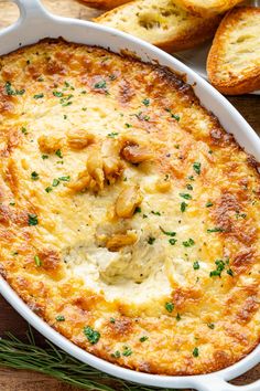 an oval casserole dish filled with cheese and herbs on a wooden table next to garlic bread