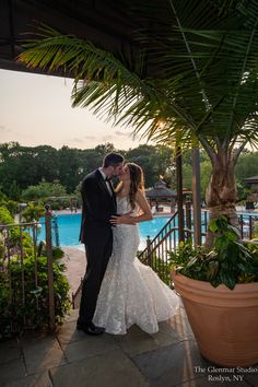 a bride and groom kissing in front of the pool at their wedding reception, surrounded by greenery