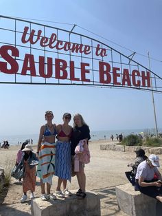 three women standing under a welcome sign at the beach