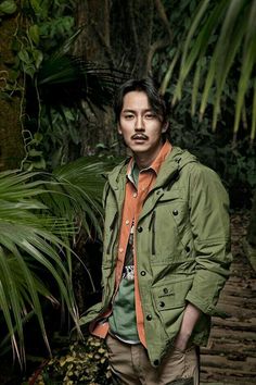 a man standing in the middle of a jungle with palm trees behind him and his hands in his pockets