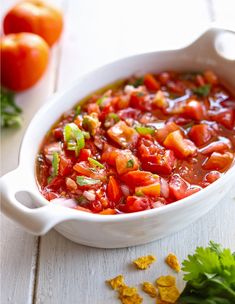 a white bowl filled with lots of food on top of a wooden table next to tomatoes