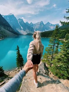 two people holding hands while standing on top of a cliff overlooking a lake and mountains