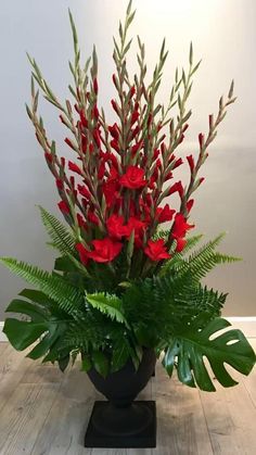 a vase filled with red flowers and greenery on top of a wooden floor next to a wall