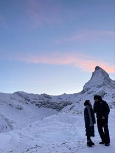 two people standing on top of a snow covered hill next to a tall mountain under a pink sky