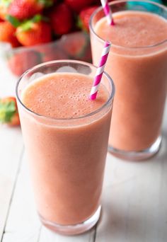 two glasses filled with smoothie sitting on top of a table next to strawberries