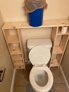 a white toilet sitting in a bathroom next to a wooden shelf with a blue trash can